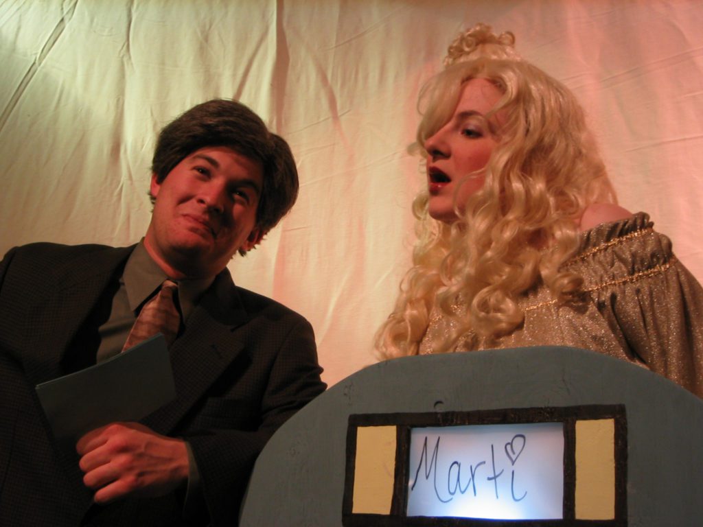 A male gameshow host is leaning against the podium of contestant Marti who has long blond hair and spells her name with a heart over the I.