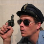 Close up on a dopey looking cop wearing sunglasses and holding his six shooter up to his face.