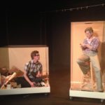 Two people talk to each other from inside plexiglass boxes. On the left is a man sitting crosslegged in a short, cluttered box. On the right is a woman sitting on a stool in a narrow box. Above is an empty clothing line.