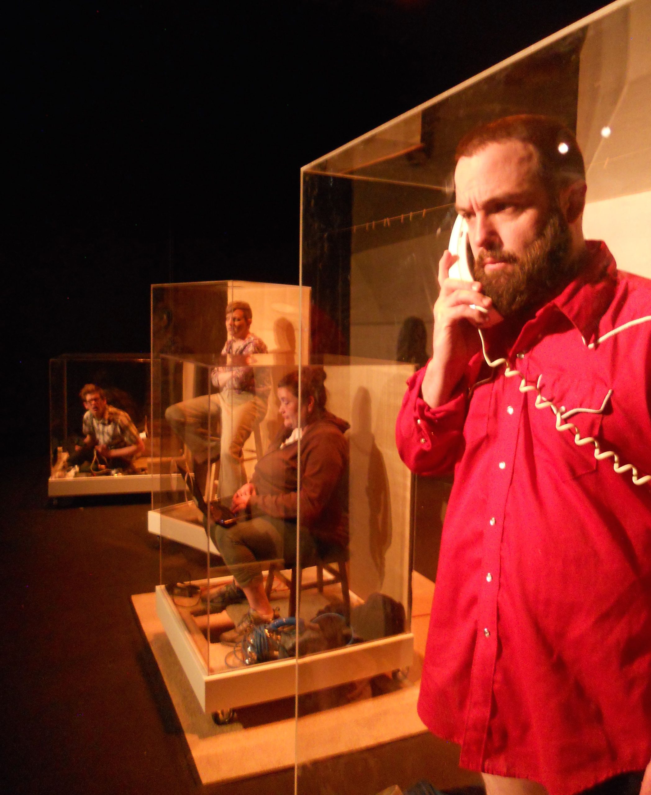 An unhappy bearded man holds a telephone behind plexiglass. In the distance are three other people sitting in large plexiglass boxes.