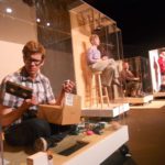 Four large plexiglass boxes are next to each other. In the closest one, a man sits cross-legged looking through a small cardboard box. He is confused. There is a person in each box in the distance.