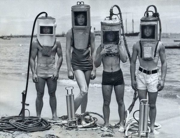 A vintage photo of 4 people in early style SCUBA gear stand in a row on a beach.