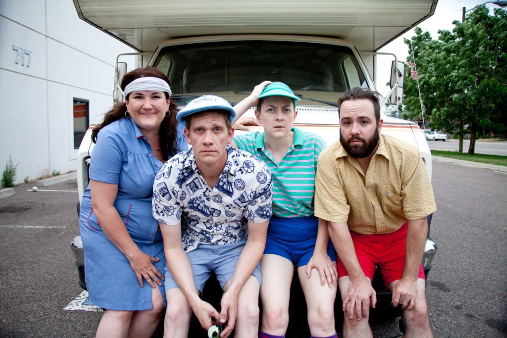 Four people wearing 70s-inspired clothes sit on the bumper of an old camper van. They all lean in towards the camera, three with serious faces, one smiling.