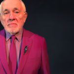 an older bearded man in a bright fuchsia suit looks sternly at the camera. He is surrounded by darkness and has a lightning bolt lapel pin.