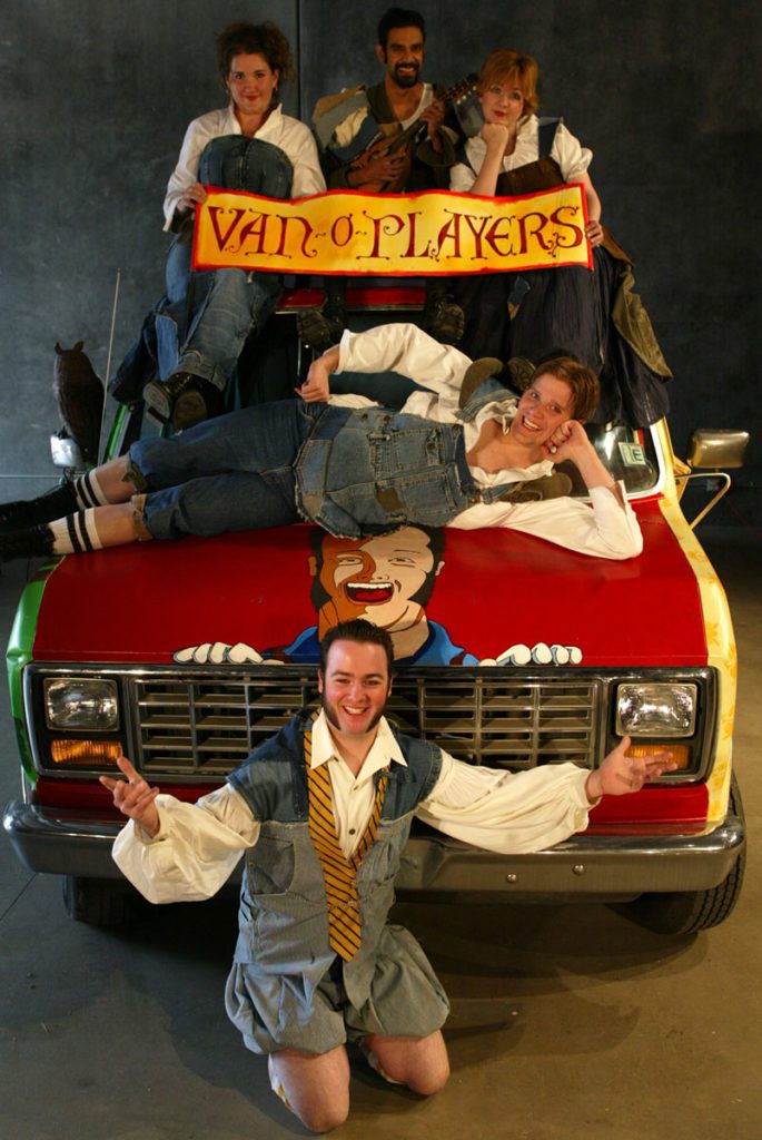 Five people are draped on and around a painted van. They all wear makeshift Shakespearean clothing. In front is a smiling man with his hands out. The hood of the van has a large smiling portrait of him. One smiling man is draped across the hood. Sitting on top of the van are three more people holding a banner that says “van-o-players”.