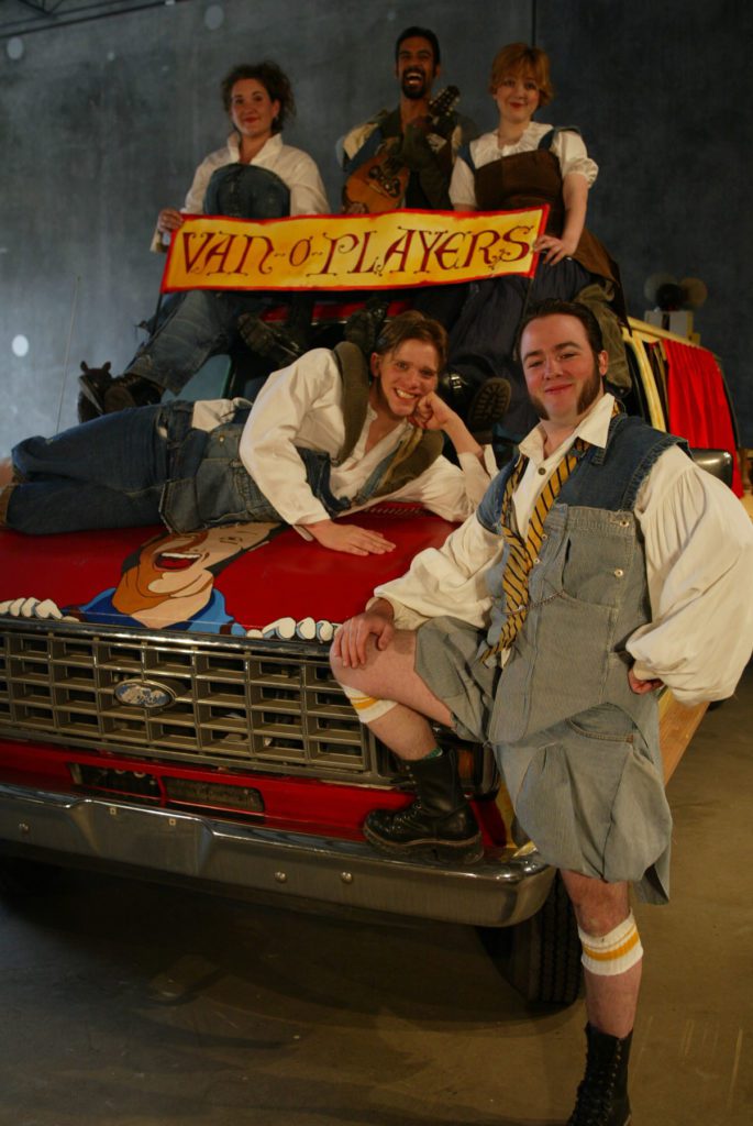 Five people are draped on and around a painted van. They all wear makeshift Shakespearean clothing. In front is a smiling man with his leg up on the bumper. The hood of the van has a large smiling portrait of him. One smiling man is draped across the hood. Sitting on top of the van are three more people holding a banner that says “van-o-players”.