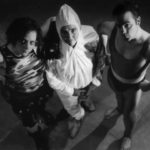 Three people are posed in outrageous costumes looking up at the camera. One woman wears Mad Max style armor and a camouflage tank top. One woman is dressed in a hazmat suit. The man is dressed in super hero attire.