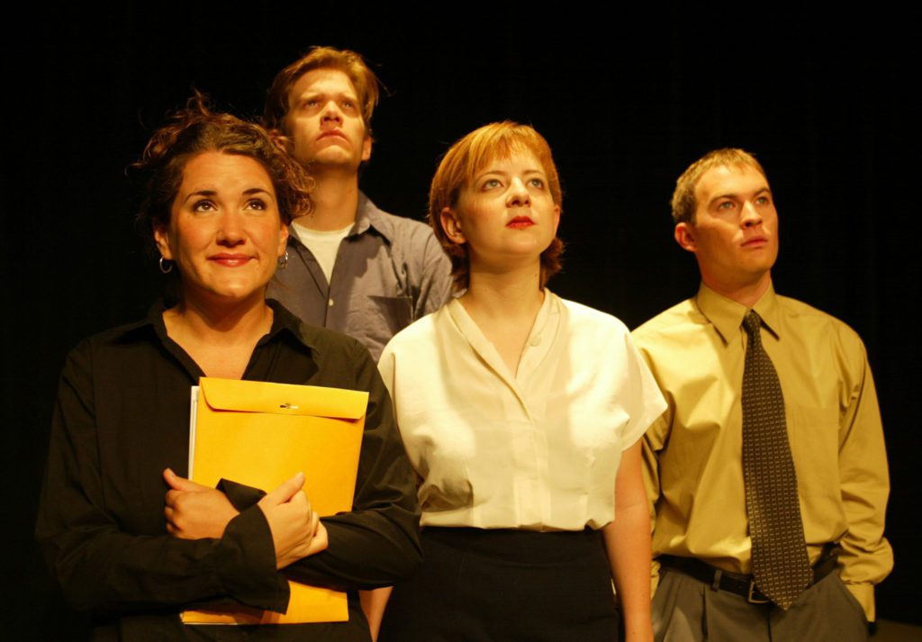 Four people wearing business casual attire stand together. All of them are looking up. One is holding a large yellow envelope.