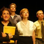 Four people wearing business casual attire stand together. All of them are looking up. One is holding a large yellow envelope.