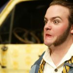Close-up of a man with large sideburns. He is bug-eyed and grimacing. In the background, out of focus, in the front of a yellow van.