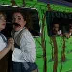 Two women clasp hands as they look out, wide-eyed. One of the women wears a fake mustache. Behind them, peaking out of the window of a van that has been painted like a forest, three men are watching.