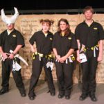 Four confused people wearing utility suits and utility belts with origami paper props hanging off of them are standing in a line. One of the people is wearing a huge Viking horned helmet made of paper. The helmet is covered in the word 'helmet.'