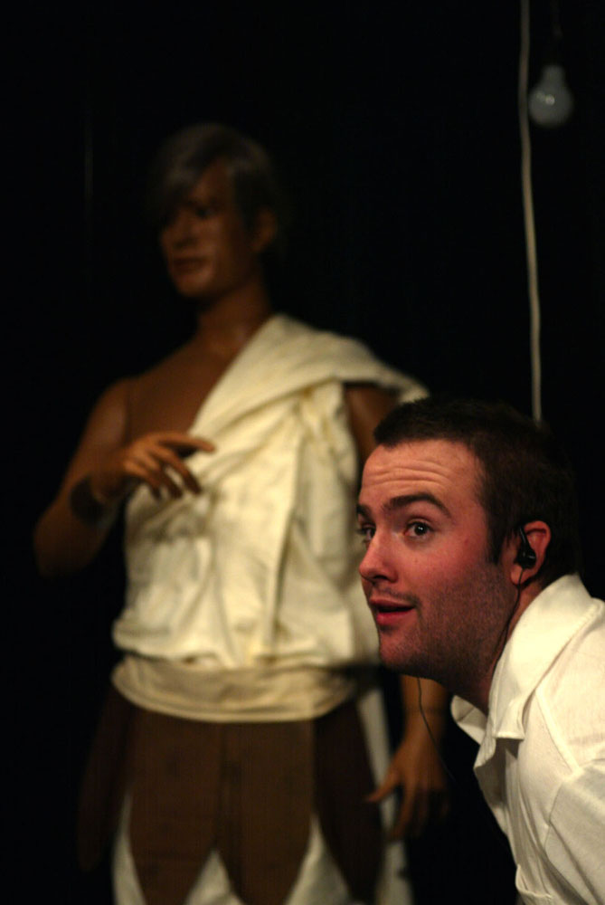 A man wearing black earphones and a white polo with a large logo on the pocket stands in front of a mannequin in a white toga