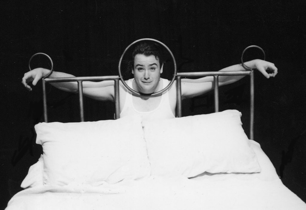 A man is pictured standing behind the headboard of a large bed. The man has positioned himself with hands and head pushed through openings in the headboard to make it seem like he’s been locked in a stockade.