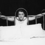 A man is pictured standing behind the headboard of a large bed. The man has positioned himself with hands and head pushed through openings in the headboard to make it seem like he’s been locked in a stockade.