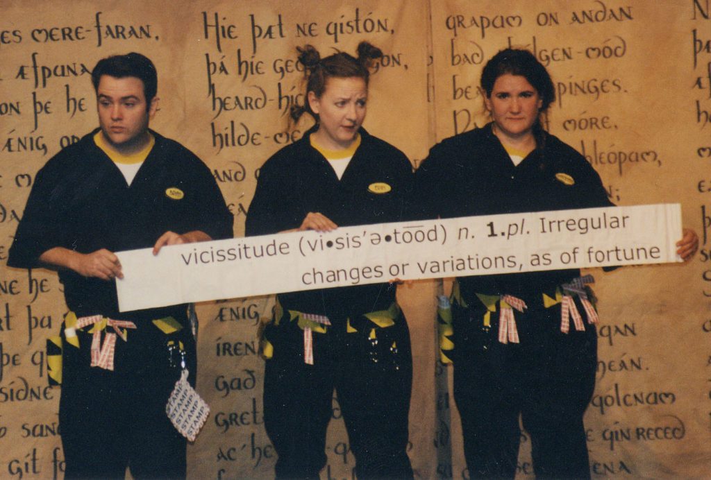 Three people stand in front of a backdrop with the Old English Poem Beowulf written on it. They are wearing utility suits with utility belts that have origami props hanging off of them. They are holding up a paper banner that shows the definition of the word vicissitudes.