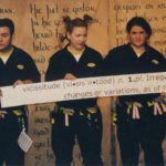 Three people stand in front of a backdrop with the Old English Poem Beowulf written on it. They are wearing utility suits with utility belts that have origami props hanging off of them. They are holding up a paper banner that shows the definition of the word vicissitudes.