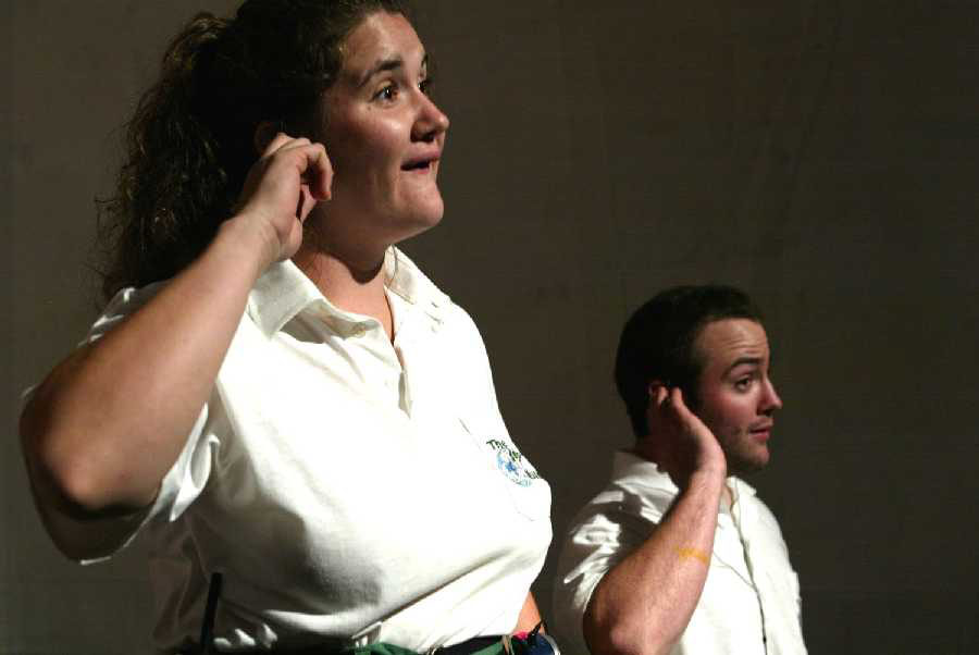 Two people in white polo shirts stand with elbows out. The man on the right wears black earphones and points to the walkman on his hip.