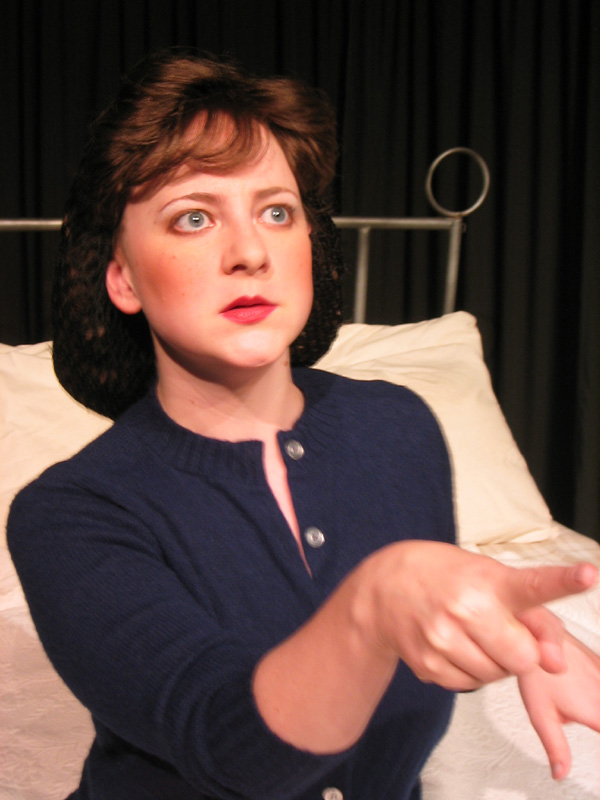 Close up of a woman sitting on a large bed wearing a blue button up sweater. Her long hair is gathered up in a hairnet 1940s style. She is pointing at something in the distance.