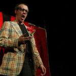A tall man wearing black rimmed glasses and a plaid blazer with an oversized flower boutonnière lectures in front of a red curtain backdrop.