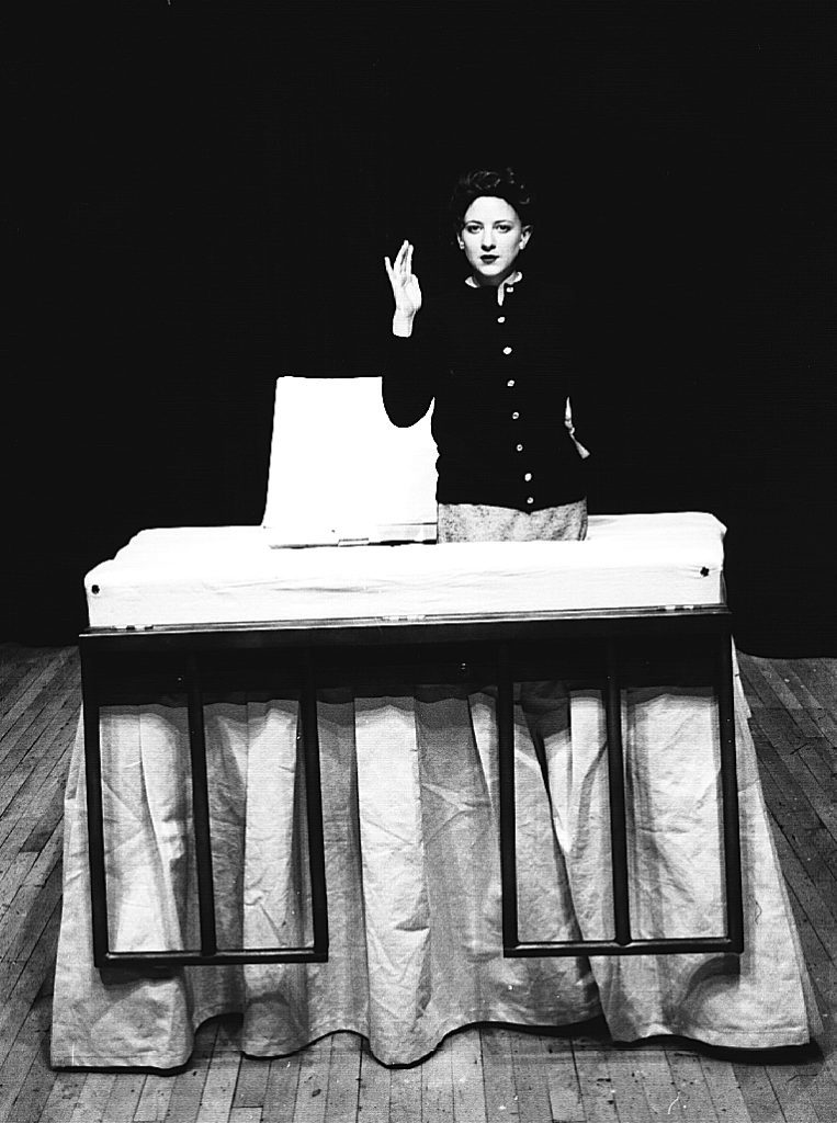 A trapdoor has been cut into a giant bed. A woman is coming out of the trap door and seems to be waving at the camera.