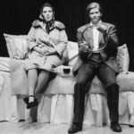 A man and a woman are seated on the side of a large bed that’s been made to look like the interior of an old car. The man holds a circular part of the headboard like a steering wheel.