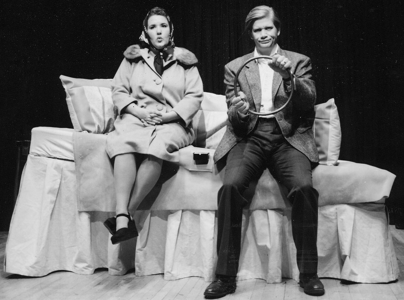 A man and a woman are seated on the side of a large bed that’s been made to look like the interior of an old car. The man holds a circular part of the headboard like a steering wheel.