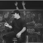 A student sits in front of a large chalkboard. The class seated behind him is drawn in chalk. The chalk students have real arms that poke out from behind the chalkboard.