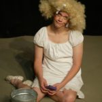 A very tall and homely Cinderella sits on the floor looking forlorn with a huge blond curly wig and a mop bucket and sponge.