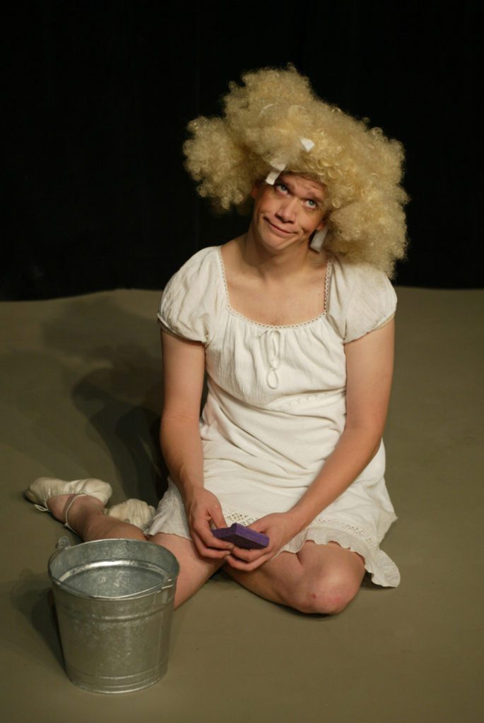 A very tall and homely Cinderella sits on the floor looking forlorn with a huge blond curly wig and a mop bucket and sponge.