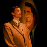 A man in a brown suit stands looking thoughtfully up and to his left. He is standing in front of a large mural of a man’s face with dark hair.