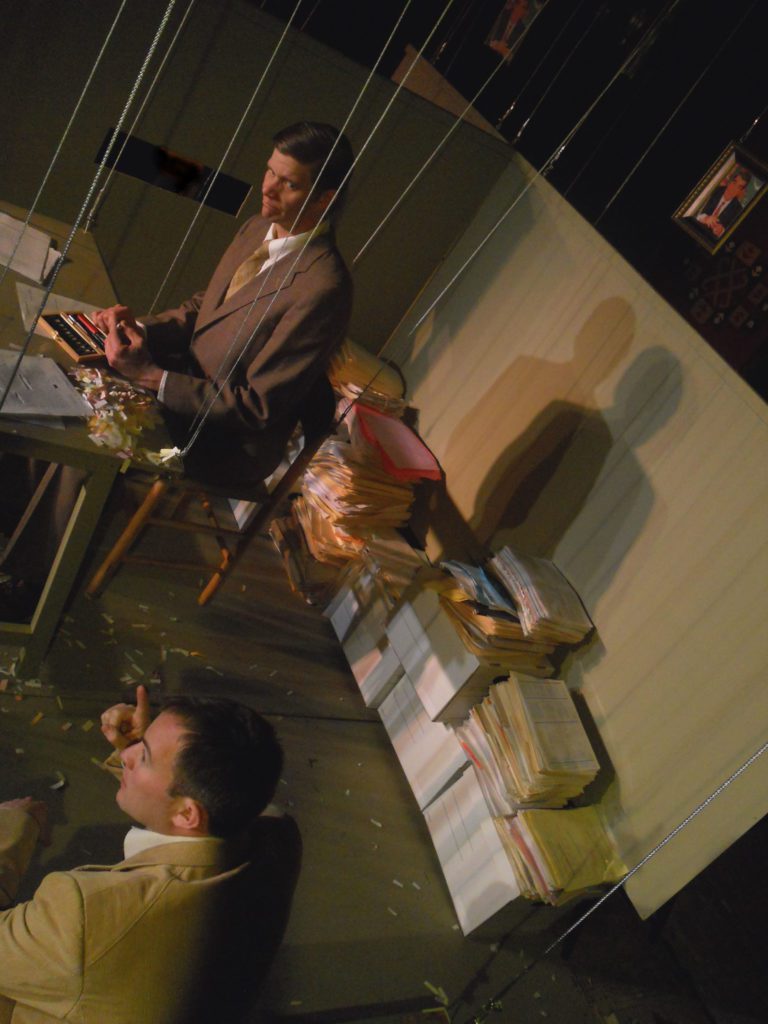 A down-shot of a room with several large stacks of paper piled up on the floor. A man in a brown suit, sitting at an awkwardly small desk that is suspended above the floor by wires, is holding a cutting blade. The man in the suit is looking concerned at another man in the foreground in a tan jacket and light green shirt who is speaking.