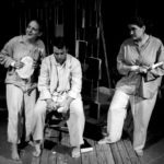 A man sits dejected peeling a potato. Behind him books hang from pieces of rope. Two women stand beside him dressed in rags and wearing cuffs made of plaster.