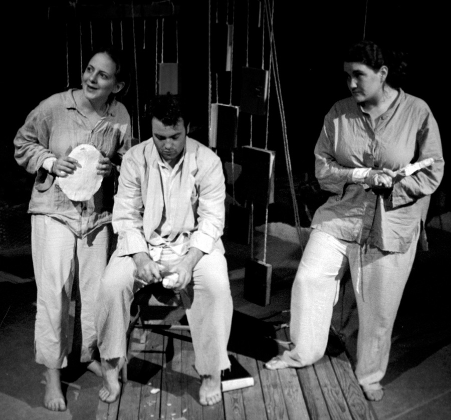 A man sits dejected peeling a potato. Behind him books hang from pieces of rope. Two women stand beside him dressed in rags and wearing cuffs made of plaster.