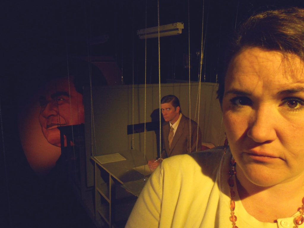 In the foreground is a woman’s face looking forward and frowning. In the distance a man sits looking concerned. He is seated at a very small desk that is suspended by wires to the ceiling. Behind the man is a large mural portrait of an imposing man on the wall.