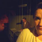 In the foreground is a woman’s face looking forward and frowning. In the distance a man sits looking concerned. He is seated at a very small desk that is suspended by wires to the ceiling. Behind the man is a large mural portrait of an imposing man on the wall.