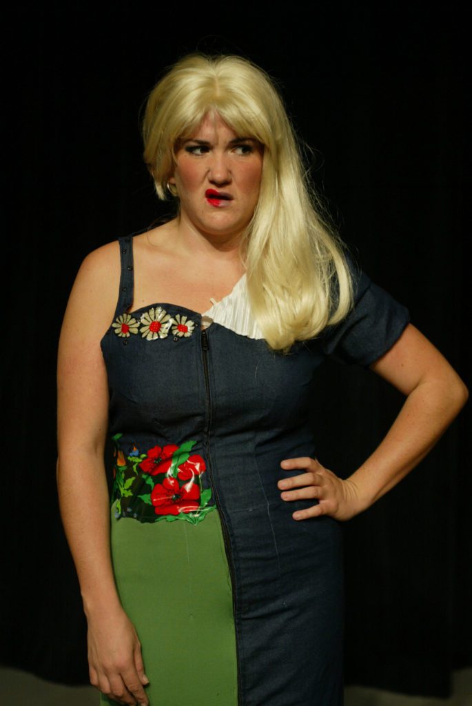 A woman looks annoyed. She is divided in half. One side of her hair is short, the other is long. One half of her dress has flowers, the other has a large white collar. only half of her mouth has lipstick. She plays both of the stepsisters.