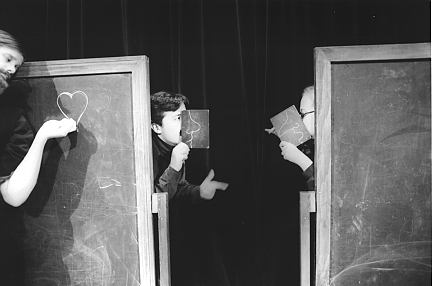 Two large, rolling chalkboards have been pushed together. A man and a woman peek out from the middle of the two chalkboards in profile. The man and the woman wear chalkboard masks that make their noses seem large and fanciful. Another man with a large goatee stands on the far left. He leans in to lovingly draw a heart on one of the chalkboards.
