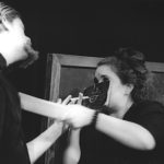 Close up of a man with a large goatee trying to speak with a woman who is wearing a small chalkboard mask. Drawn on the chalkboard mask is a nose and mouth. The woman seems to be smoking a long piece of chalk like a cigarette and is pushing the goateed man back with a large eraser.