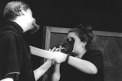 Close up of a man with a large goatee trying to speak with a woman who is wearing a small chalkboard mask. Drawn on the chalkboard mask is a nose and mouth. The woman seems to be smoking a long piece of chalk like a cigarette and is pushing the goateed man back with a large eraser.