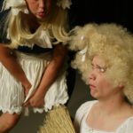 A woman in blond pigtails leans over blowing a raspberry in the face of Cinderella, who looks upset and holds a broom.