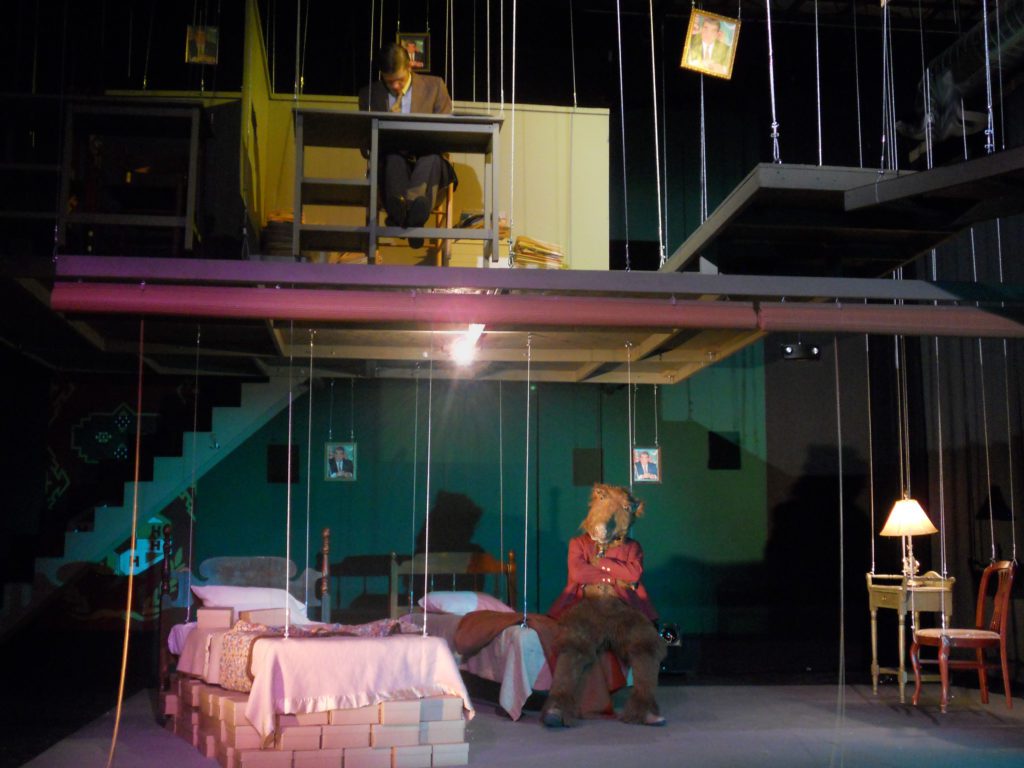 There are two rooms one directly above the other in which all furniture and walls in both rooms are suspended by wires. In the top room a man leans over a small desk. To his left is a walkway supported by wires. In the room below a person in a camel costume sits on the front edge of a wire-suspended bed. The room is cluttered with boxes.