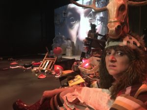 On the far right a person looks unhappily at the camera. They have a large headpiece that includes a plastic horse head with antlers. In the background, the floor is littered with crud. Projected on the wall is an extreme close-up of someone's face.