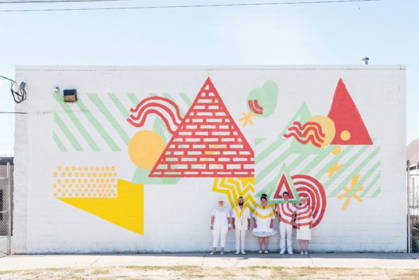 Five people stand in front of a large white wall of colorful graffitied shapes. The people, all wearing white, are painted to blend in with the graffiti.