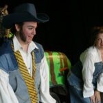 A man with sideburns and a large cowboy hat is talking. Over his shoulder is a plastic owl staring at the camera. A woman standing with her hands on her knees is smiling at the man.