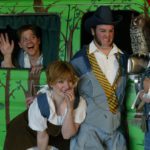 Three people stand in front of a van that has been painted to look like a forest, complete with a fake owl on the side mirror. The people are dressed in homemade Shakespearean outfits and look concerned. Two additional people peak out from the van's window.
