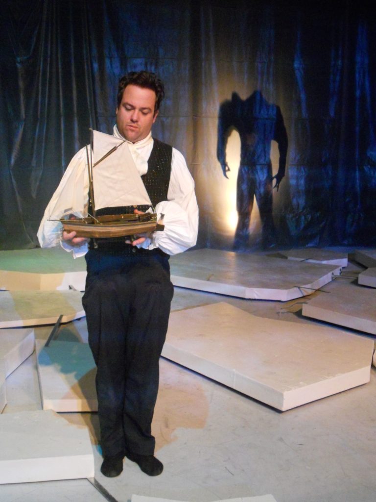 Percy Shelley stands in the middle of a sea of icebergs holding a small sailboat while in the background the shadow of a large monster looms over him.