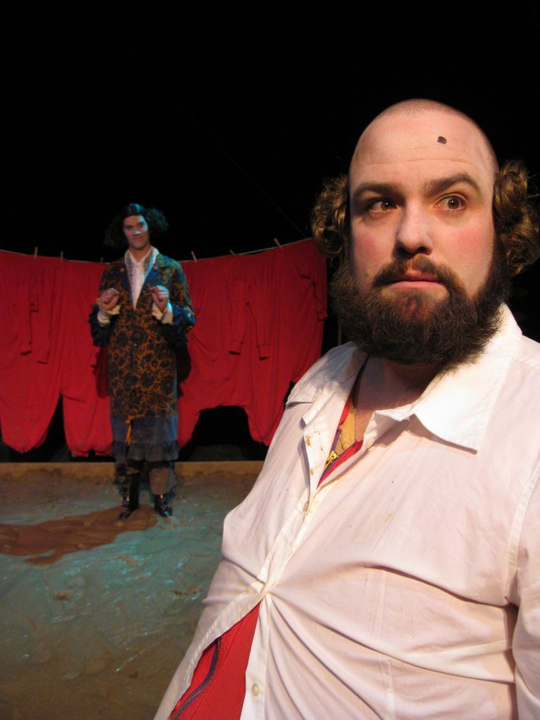 In the foreground, a large bald man with a big beard and beauty mark on his forehead stares out. Behind him, a man in a colorful jacket stands in a shallow mud pit with a laundry line of red long johns behind him.