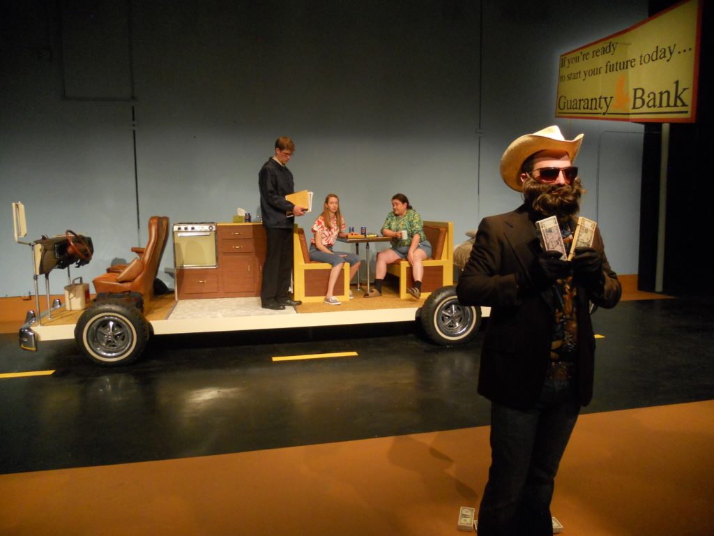 A man with a fake beard and sunglasses holds piles of cash. Behind him two people sit in an RV without walls. Next to them stands an FBI officer holding some files.