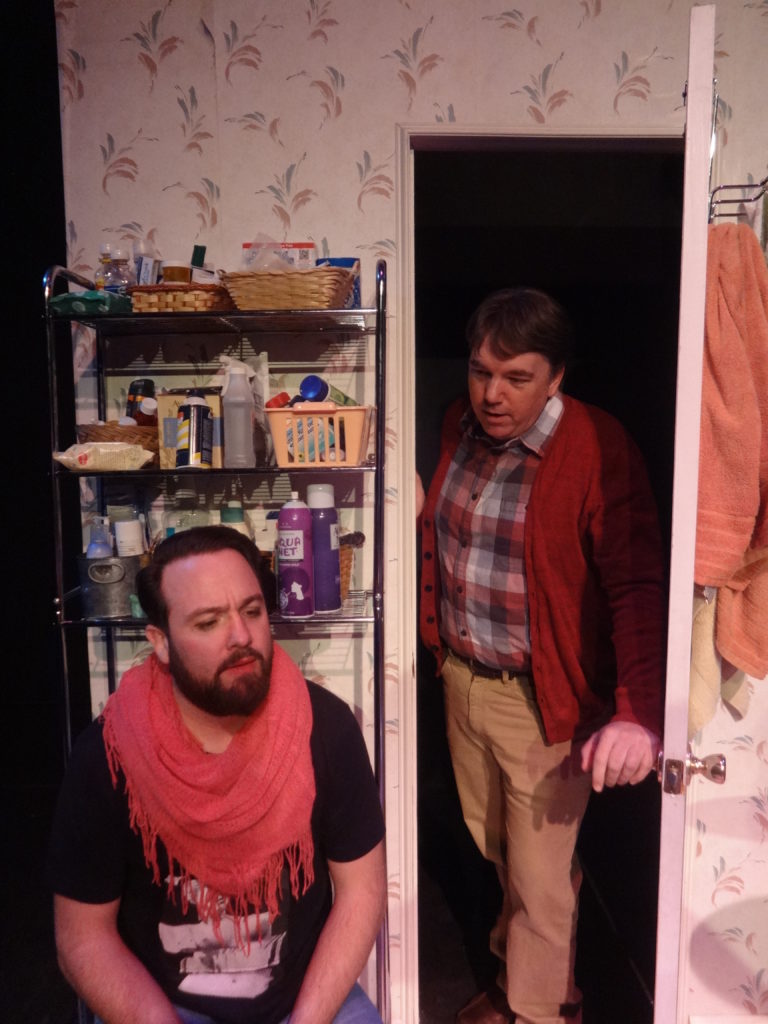 A bearded person sits on the toilet in a bathroom crowded with beauty products. Another person opens the door to check on them.